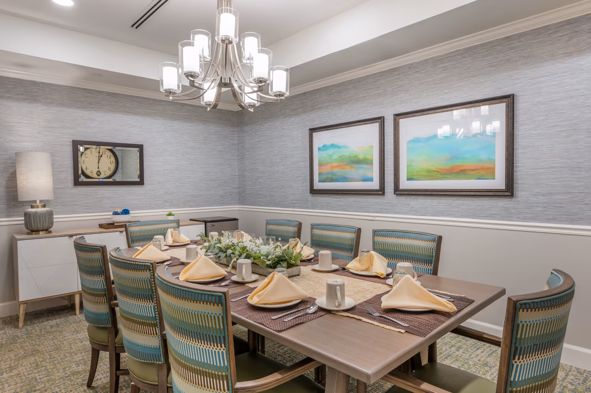 A dining room with a rectangular wooden table set for eight people. Each place setting has a beige folded napkin, a white cup on a saucer, a glass, and silverware. The chairs have a patterned upholstery in shades of blue, green, and beige. On the table is a centerpiece with green foliage and candles. The room has light gray textured walls with white wainscoting, two framed colorful landscape paintings, a modern chandelier, a sideboard with a lamp, and a wall clock reflected in a mirror.