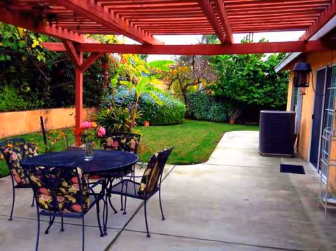 Red pergola shelters a patio with a round table, chairs, and a lush backyard.