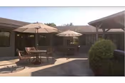 Sunny courtyard with round patio tables, chairs, and umbrellas between single-story building wings.