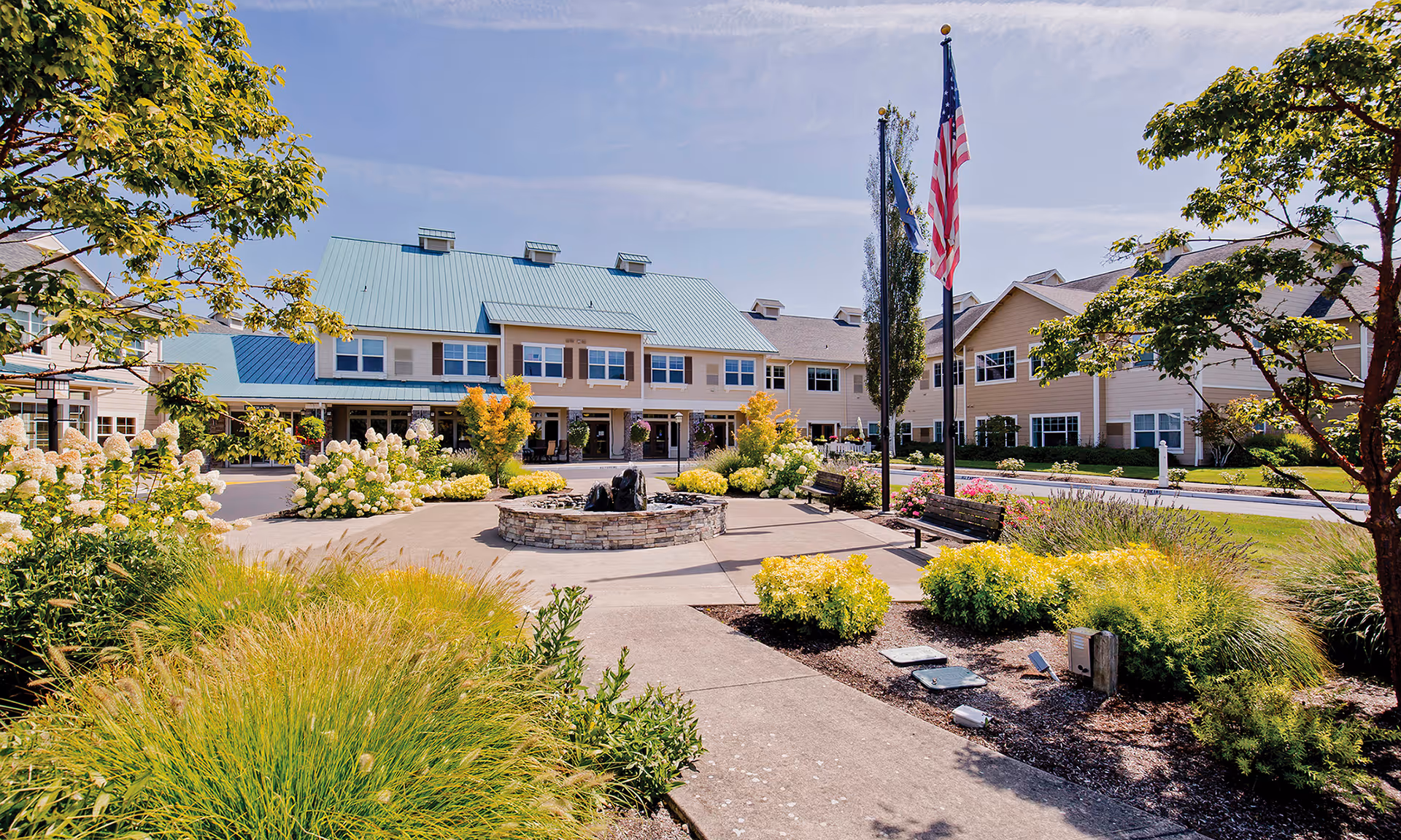 Outdoor courtyard area of a senior living facility with a circular stone fountain in the center, surrounded by benches, landscaped bushes, flowers, and trees. The building in the background has a light blue metal roof and beige siding with multiple windows. An American flag and another flag are displayed on flagpoles near the fountain under a clear blue sky.