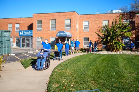 A group of people, some in wheelchairs and others standing, are outside a two-story brick building with signs that read 'Christian Care Communities' and 'Transitional Rehab & Home'. The scene is sunny with a green lawn and a paved walkway leading to the building entrance.