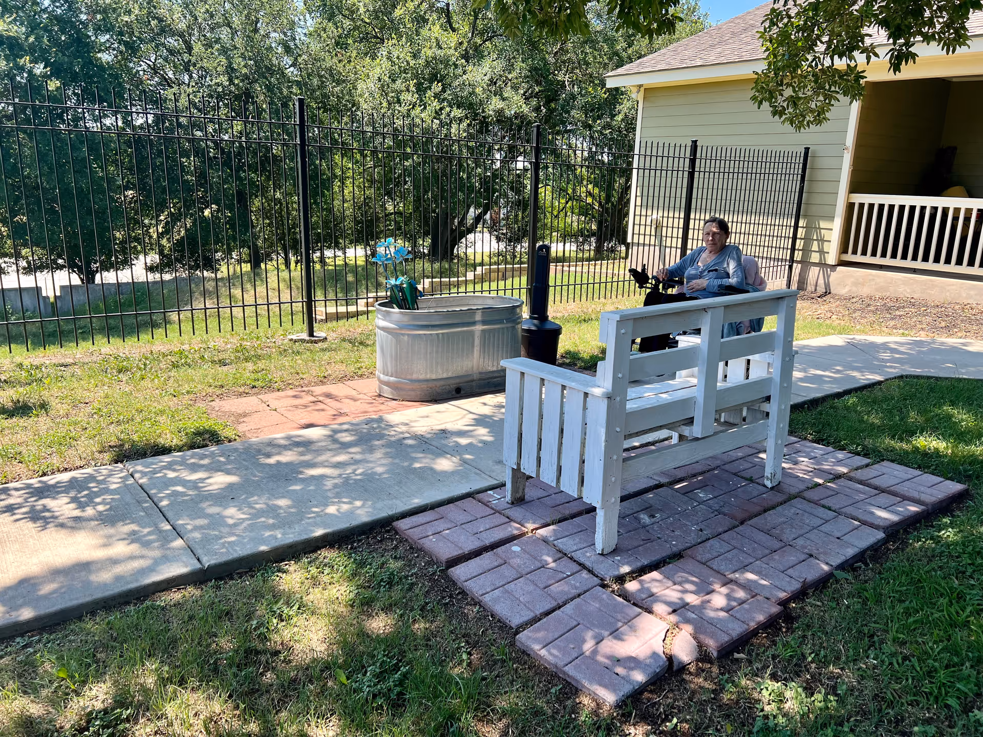An elderly person sitting on a mobility scooter near a white wooden bench on a paved patio area outside. There is a metal planter with blue decorative flowers and a black post near a black metal fence. Trees and a yellow building with a porch are visible in the background.