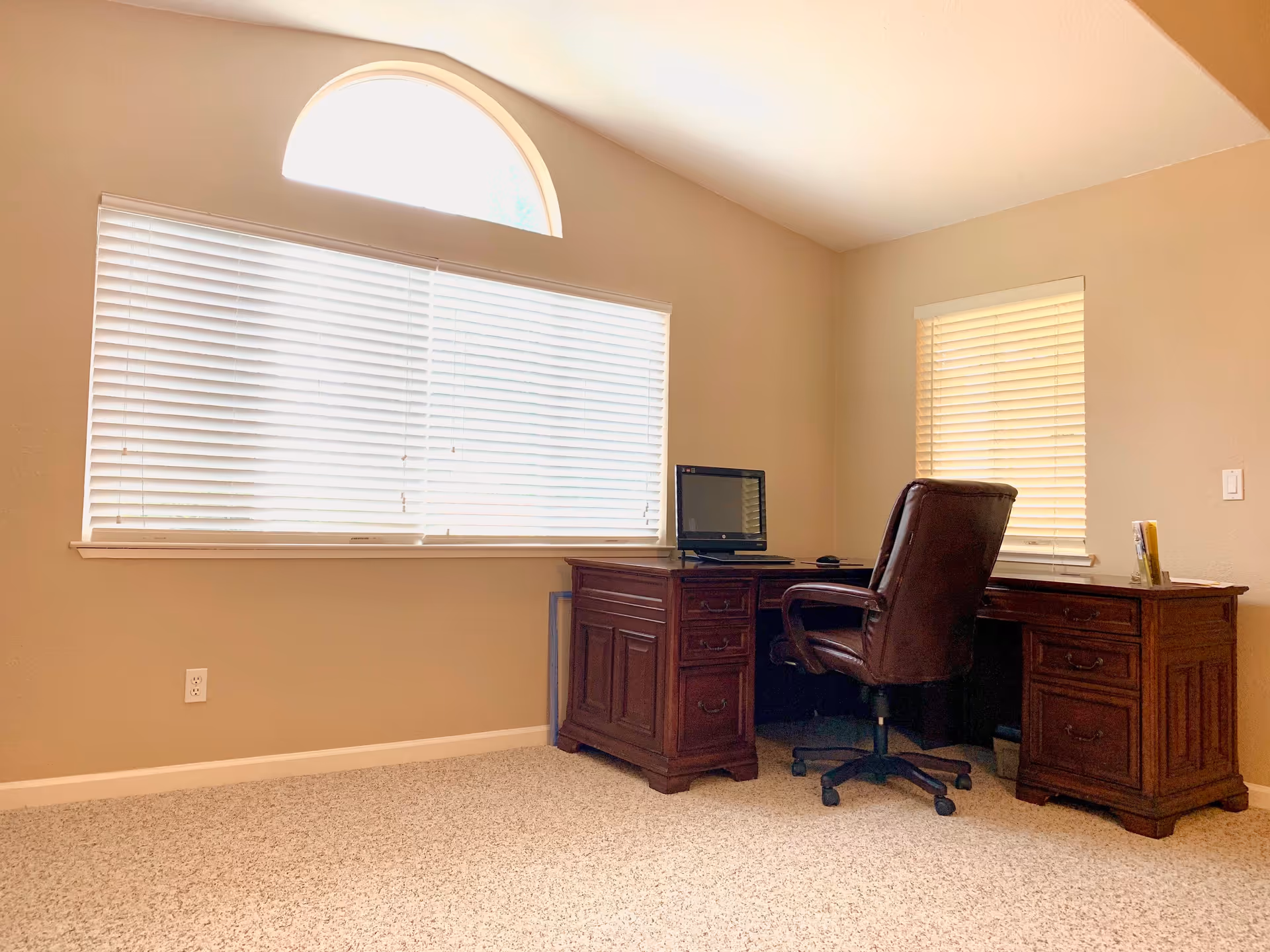 A bright room with beige walls and carpeted floor featuring a large wooden desk with multiple drawers. On the desk is a computer monitor and a brown leather office chair on wheels. The room has two windows with white blinds, one large rectangular window topped with a half-circle window, and a smaller rectangular window on the adjacent wall.