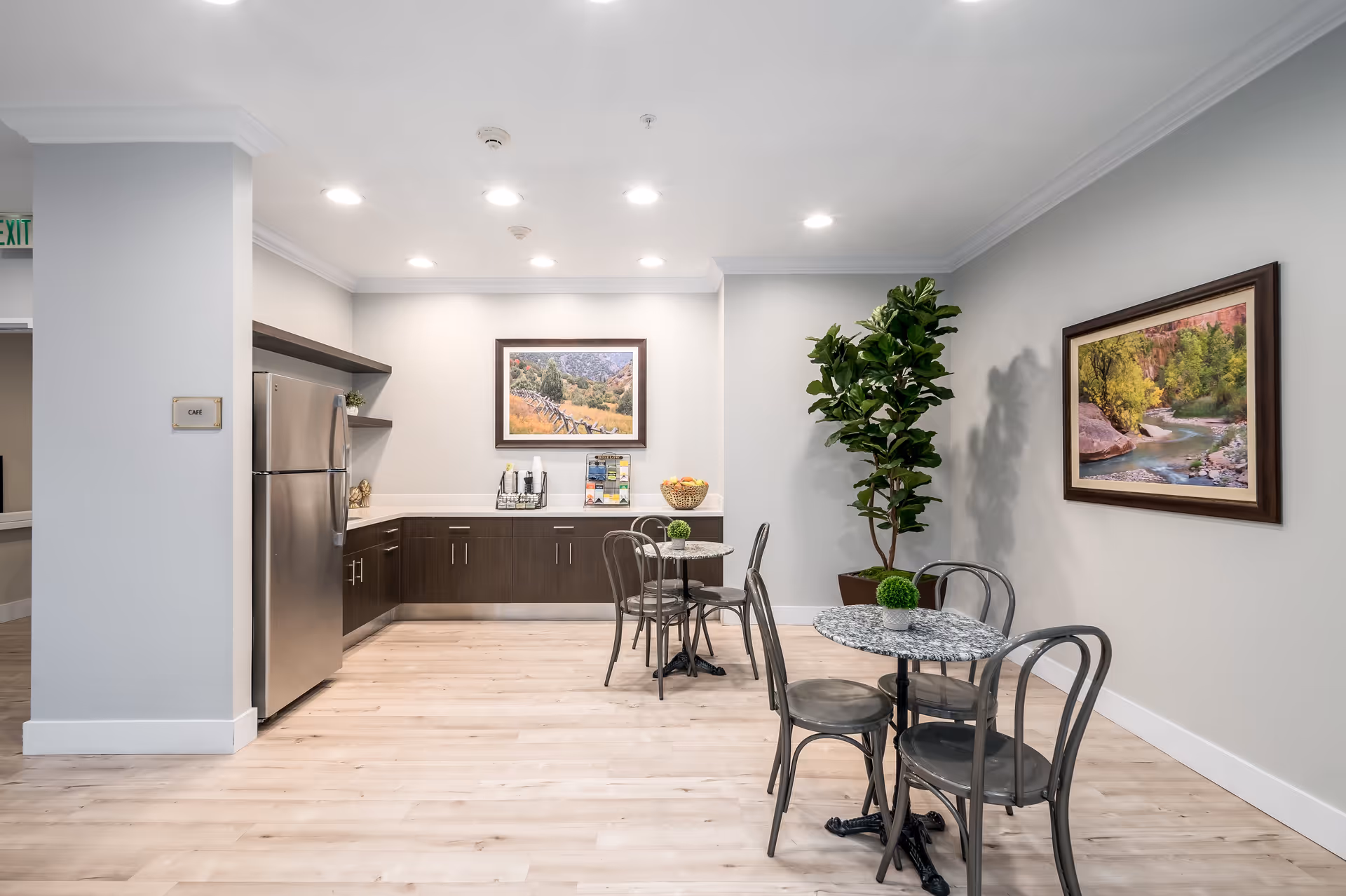 A small cafe area inside a senior living facility with two round tables and metal chairs. The room features light wood flooring, a stainless steel refrigerator, dark wood cabinets, and two framed landscape paintings on the walls. There is also a tall green potted plant in the corner and overhead recessed lighting.
