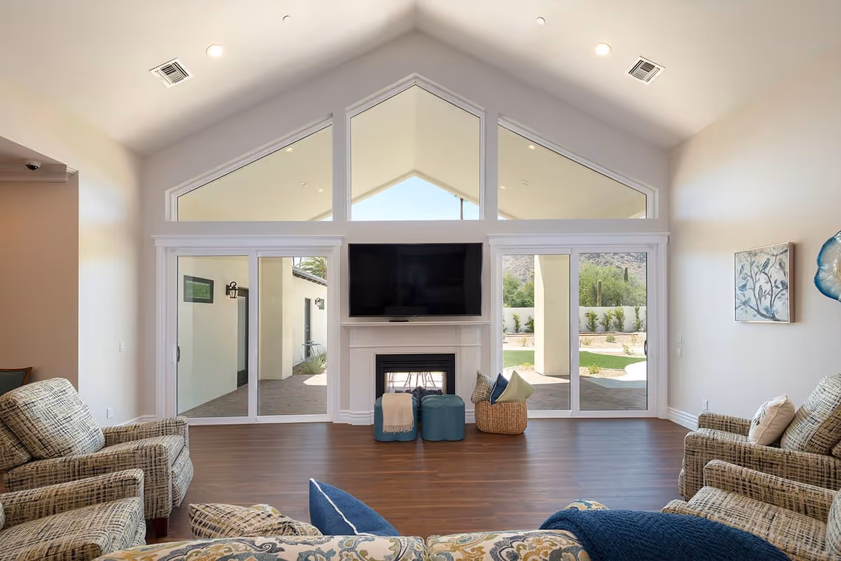 A bright living room with high vaulted ceilings and large triangular windows above sliding glass doors that open to an outdoor patio and garden area. The room features a wall-mounted flat-screen TV above a white fireplace, two blue ottomans, a basket with pillows, and patterned armchairs arranged around the space. The walls are light-colored and there is a piece of artwork on the right wall.