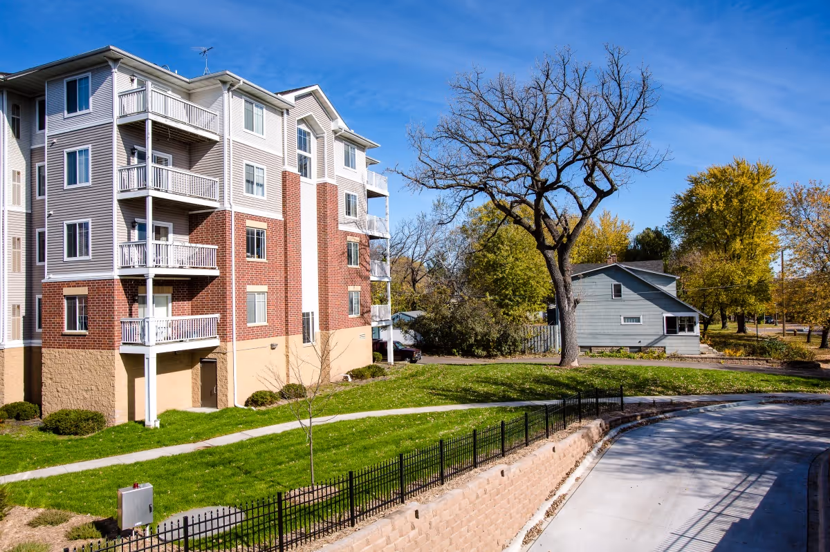 Exterior view of a multi-story residential building with balconies, surrounded by green grass, trees, and a paved driveway under a clear blue sky.
