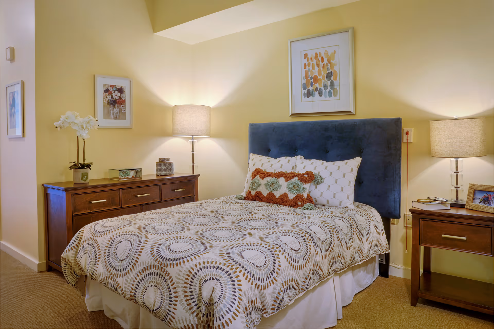 A cozy bedroom with a bed featuring a patterned bedspread and decorative pillows. The bed has a dark blue upholstered headboard. On either side of the bed are wooden nightstands with lamps, a framed picture on the wall above the headboard, and additional framed artwork on the adjacent wall. The room has soft yellow walls and carpeted flooring.