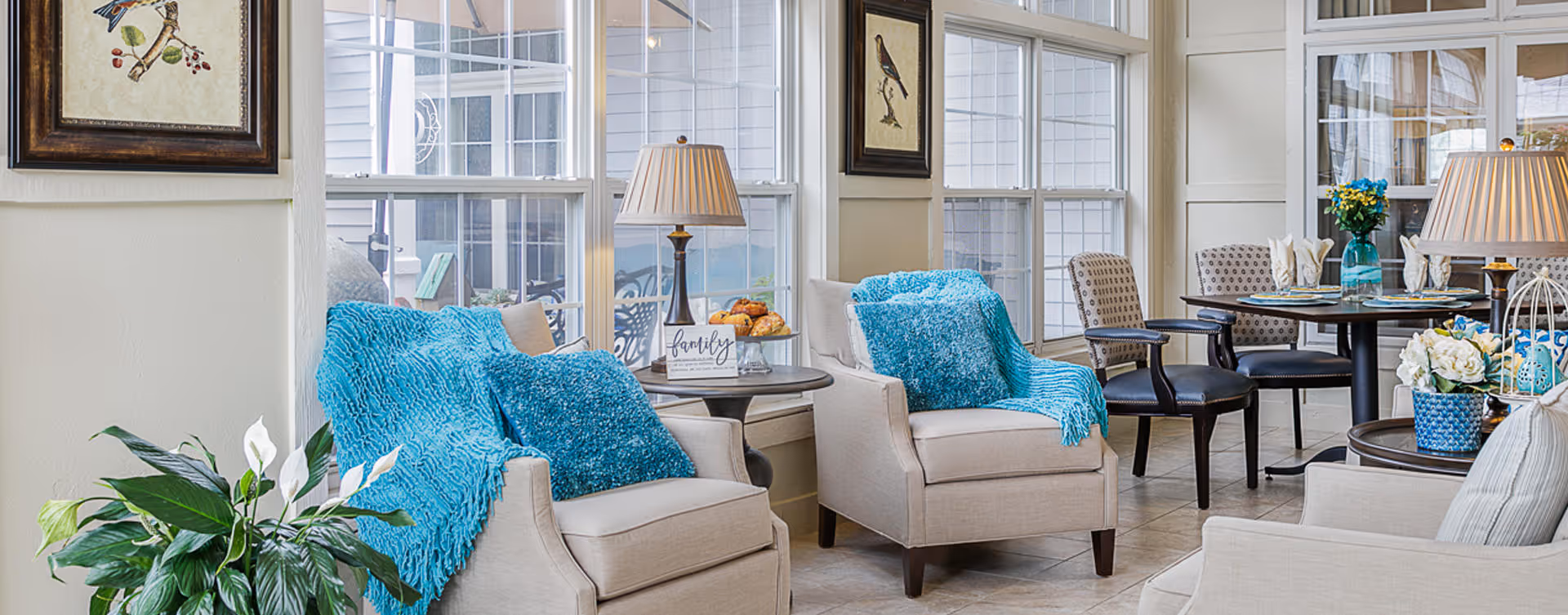 A bright and cozy living area with two beige armchairs draped with blue throws and matching pillows, a side table with a lamp and a decorative sign that says 'family', large windows letting in natural light, a round dining table set with chairs and floral centerpieces, and framed bird artwork on the walls.