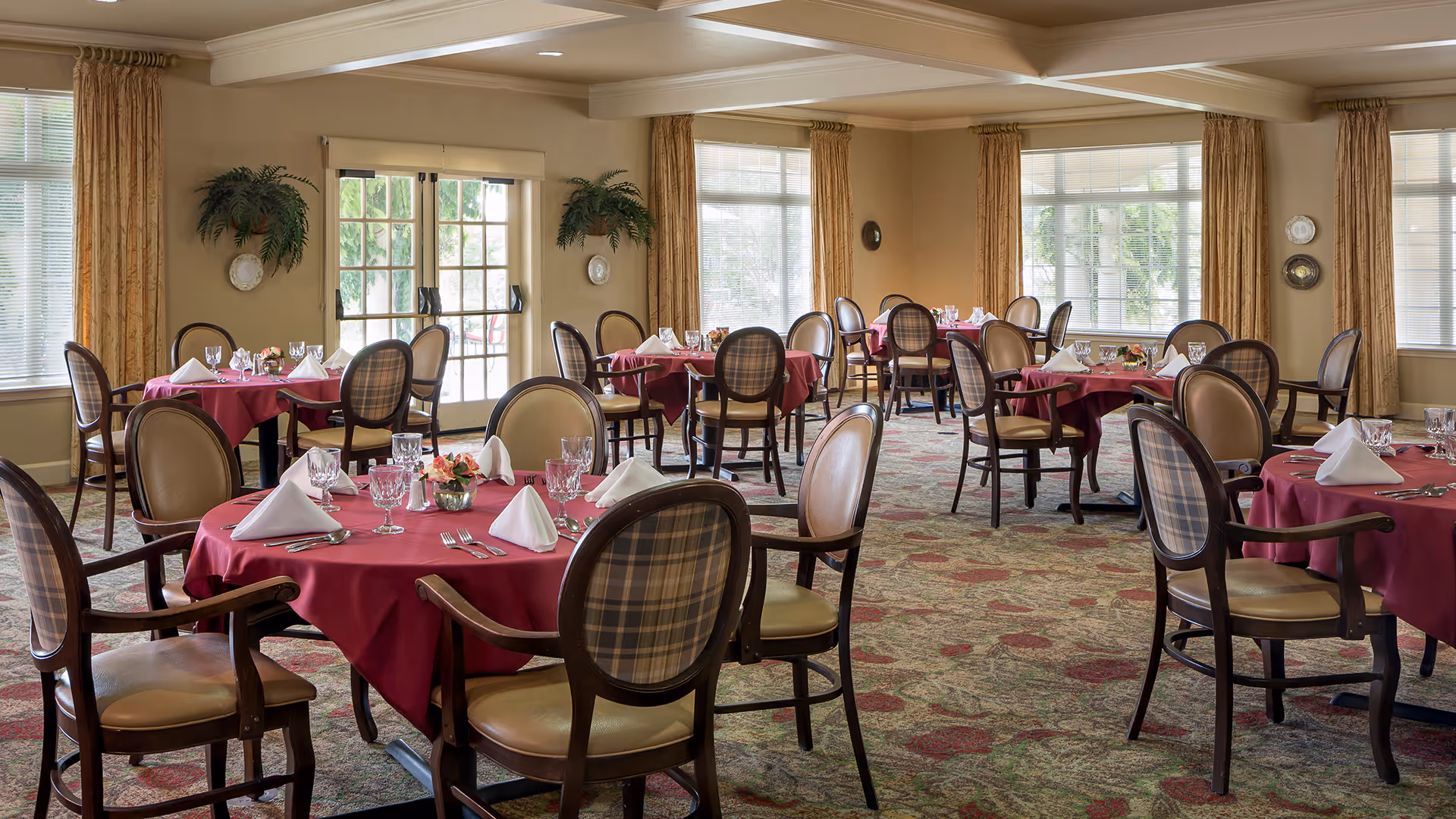 A dining room with multiple round tables covered with red tablecloths, each set with white folded napkins, glassware, and silverware. The room has large windows with beige curtains, patterned carpet, and wooden chairs with cushioned seats and backs.
