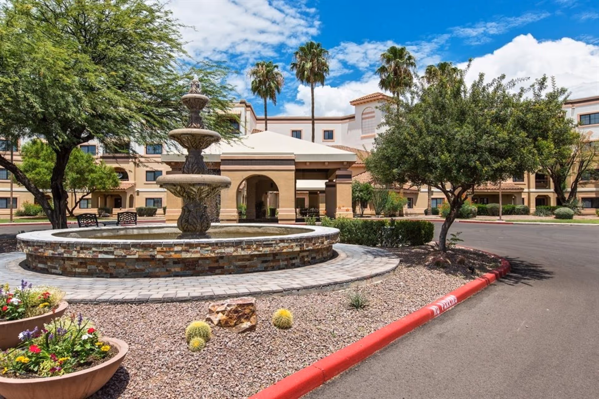 Outdoor view of The Forum at Tucson senior living facility featuring a multi-tiered stone fountain surrounded by desert landscaping with small cacti and flowering plants, palm trees, and a beige building with arched entryways under a partly cloudy blue sky.