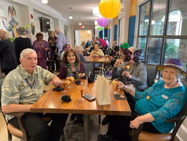A group of elderly people sitting around a wooden table in a well-lit common area decorated with colorful hanging paper lanterns. Some individuals are wearing festive masks and beads, enjoying drinks and socializing. Large windows line one side of the room, letting in natural light.