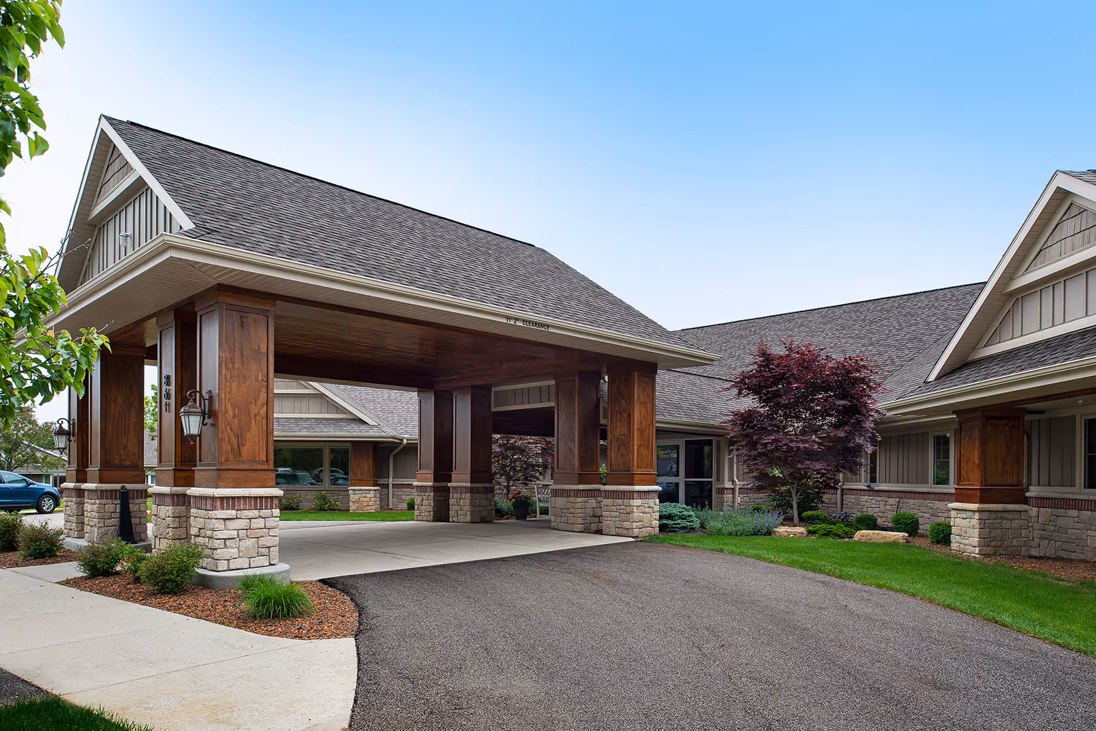 Exterior view of the American House Jenison facility showing a covered entrance with wooden pillars and stone bases, surrounded by landscaped greenery and a clear blue sky.