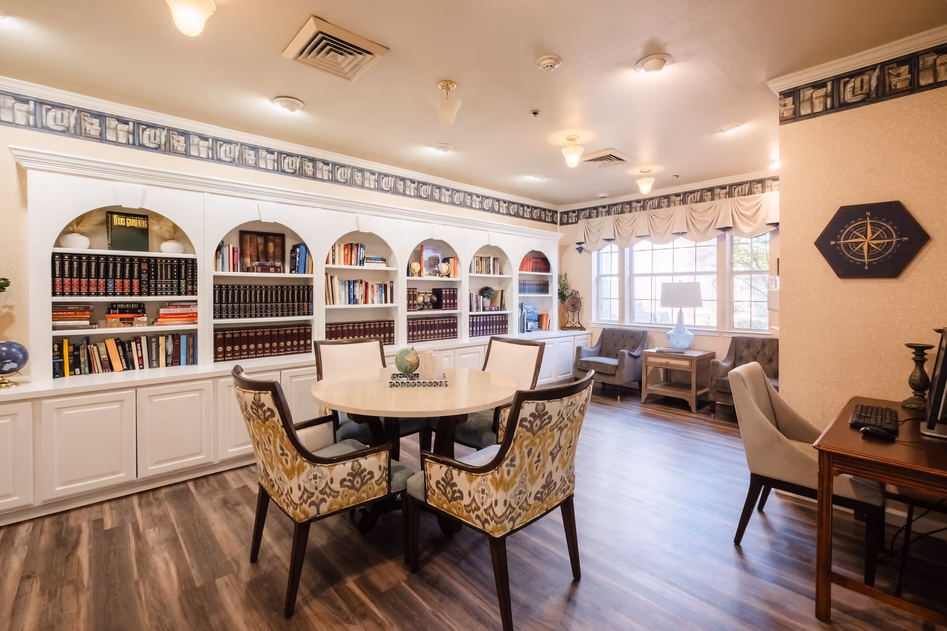 A cozy interior room with a round table surrounded by four patterned chairs in the center. Behind the table is a large built-in white bookshelf filled with books and decorative items. To the right, there are two armchairs near a window with a side table and lamp. A desk with a computer keyboard and chair is also visible. The room has wood flooring and soft lighting from ceiling fixtures.