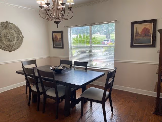 A dining room with a dark wooden rectangular table surrounded by six matching chairs with light-colored cushions. The room has wooden flooring, a window with blinds letting in natural light, two framed pictures on the walls, a decorative wall medallion, and a chandelier hanging from the ceiling.