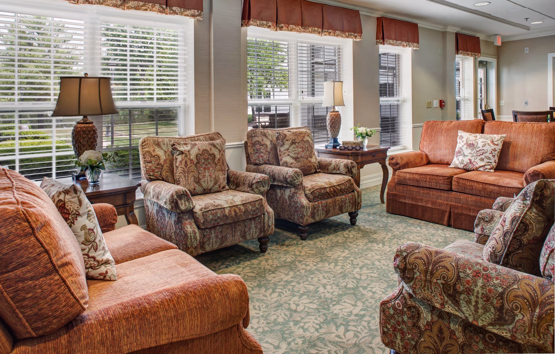 Well-lit communal living room with upholstered sofas and patterned armchairs arranged around side tables and lamps in front of large windows.