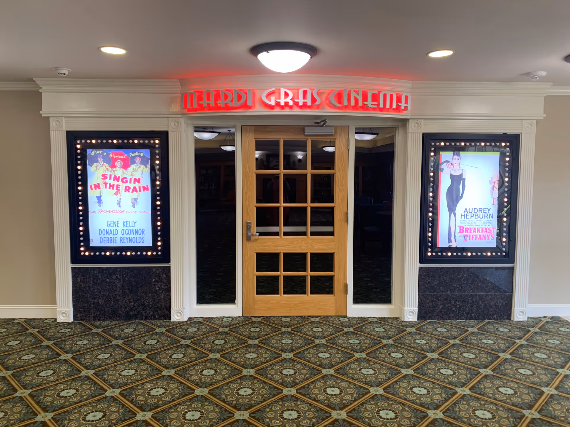 Entrance to a small cinema room named 'Mardi Gras Cinema' with a wooden door and glass panels. On either side of the door are illuminated movie posters for 'Singin' in the Rain' and 'Breakfast at Tiffany's'. The floor has a patterned carpet and the walls are light-colored with white trim.