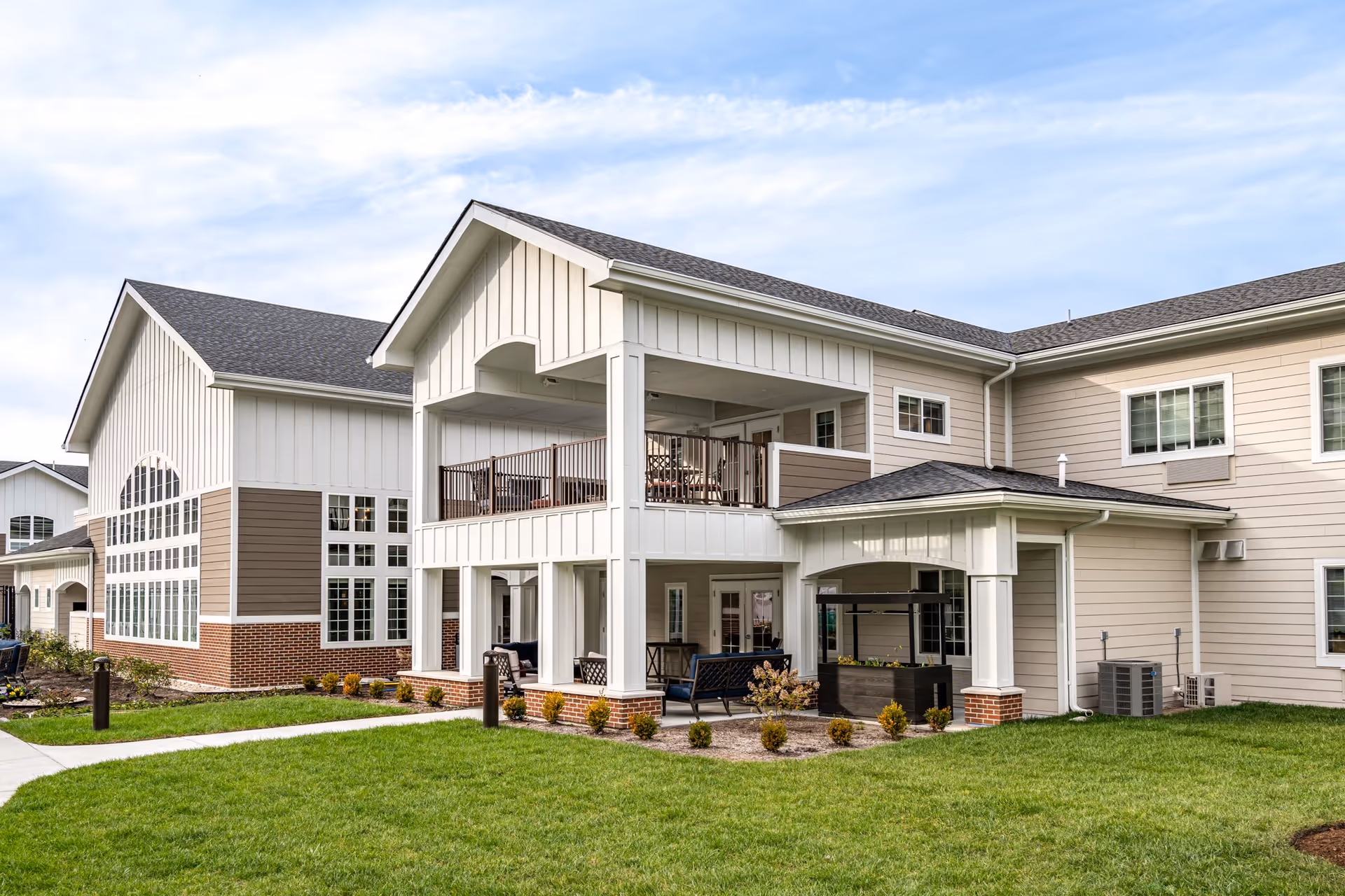 Exterior view of a senior living facility building with beige and white siding, large windows, and a covered patio area with outdoor seating. The building is surrounded by a well-maintained green lawn and small shrubs under a partly cloudy sky.