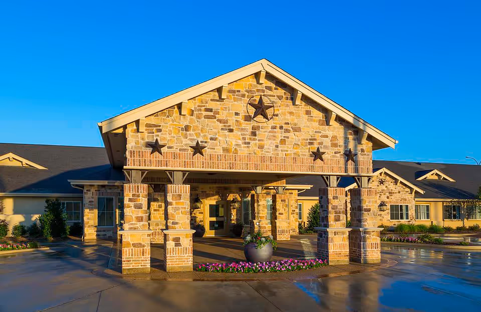 Front exterior view of Meadowbrook Memory Care Community building with stone and brick facade, decorative star accents, a covered entrance supported by stone pillars, and landscaped flower beds under a clear blue sky.