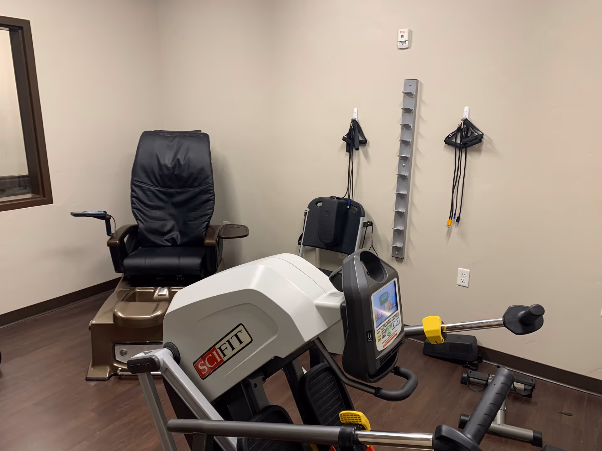 Small therapy room with an exercise machine and a black recliner chair against a beige wall.