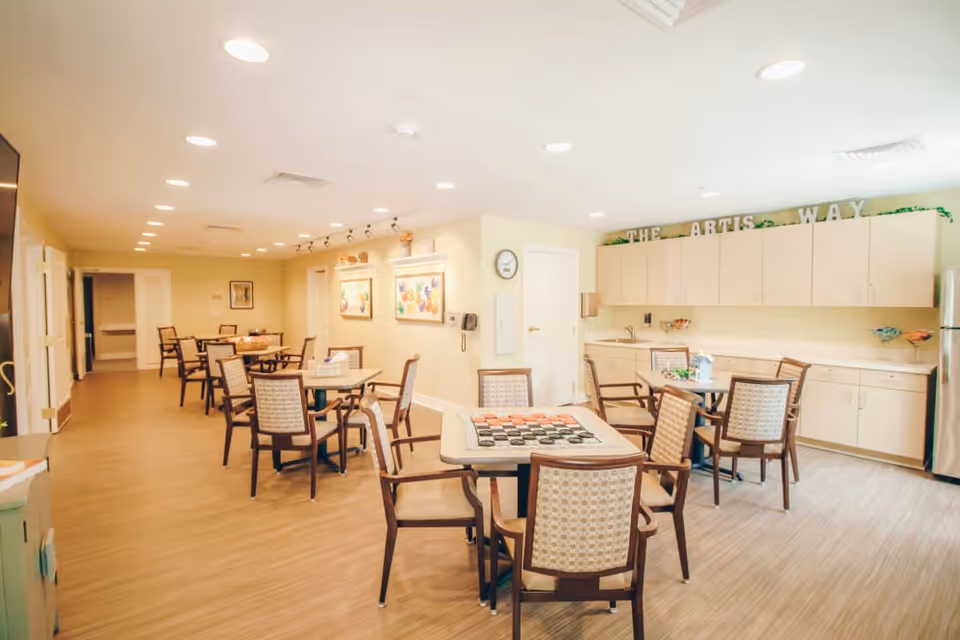 Bright communal dining and activity room with multiple tables and chairs and a kitchenette area with cabinets reading "THE ARTIS WAY".