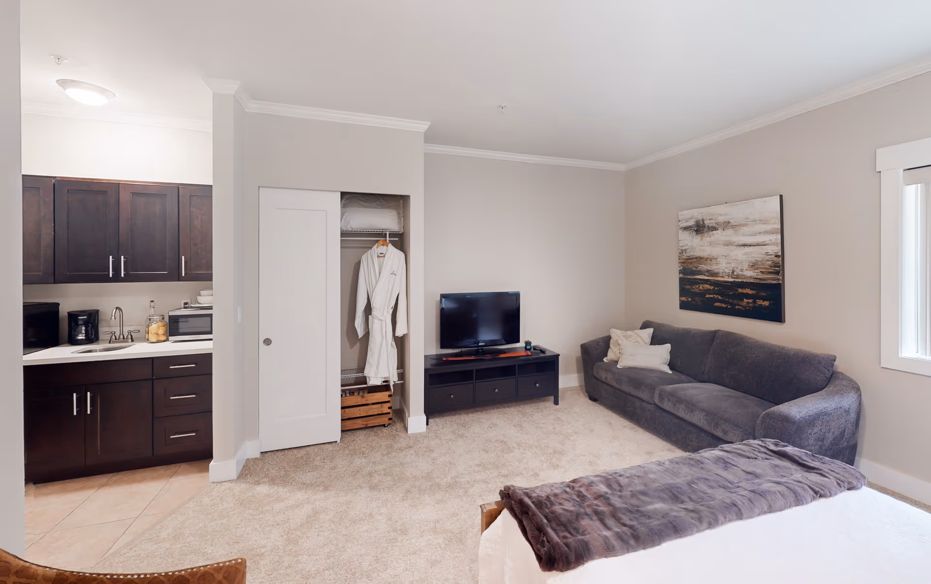 A cozy senior living room with a gray couch, a flat-screen TV on a black TV stand, a small open closet with a white robe hanging inside, and a kitchenette with dark wood cabinets and a coffee maker. The room has beige walls, carpeted floor, and a window with white trim.