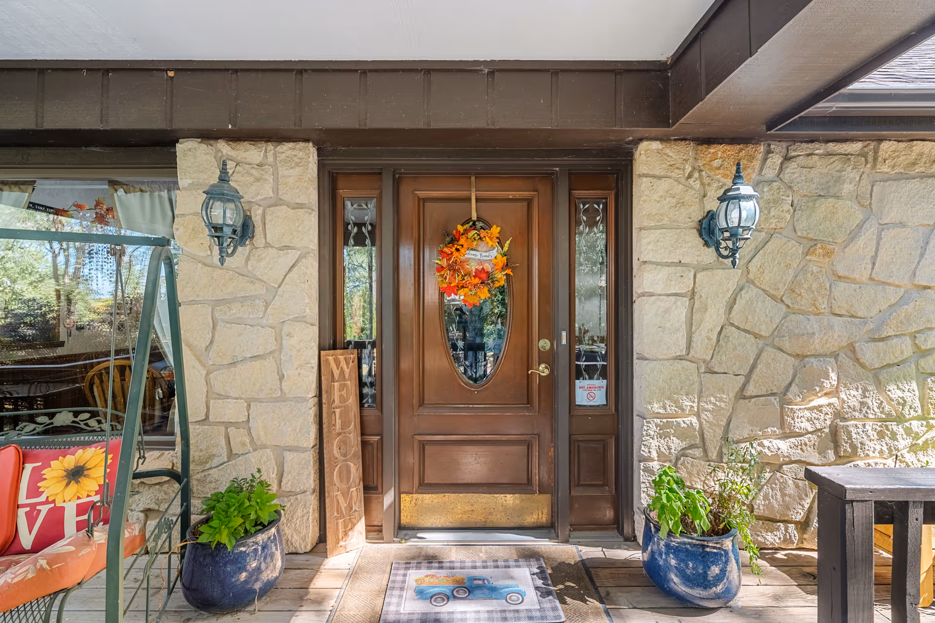 Front entrance of a building with a wooden door decorated with an autumn wreath. The door is flanked by two stone walls with outdoor lanterns mounted on each side. There are two blue ceramic pots with green plants on either side of the door, a welcome sign leaning against the left wall, and a porch swing with red and orange cushions visible on the left side.