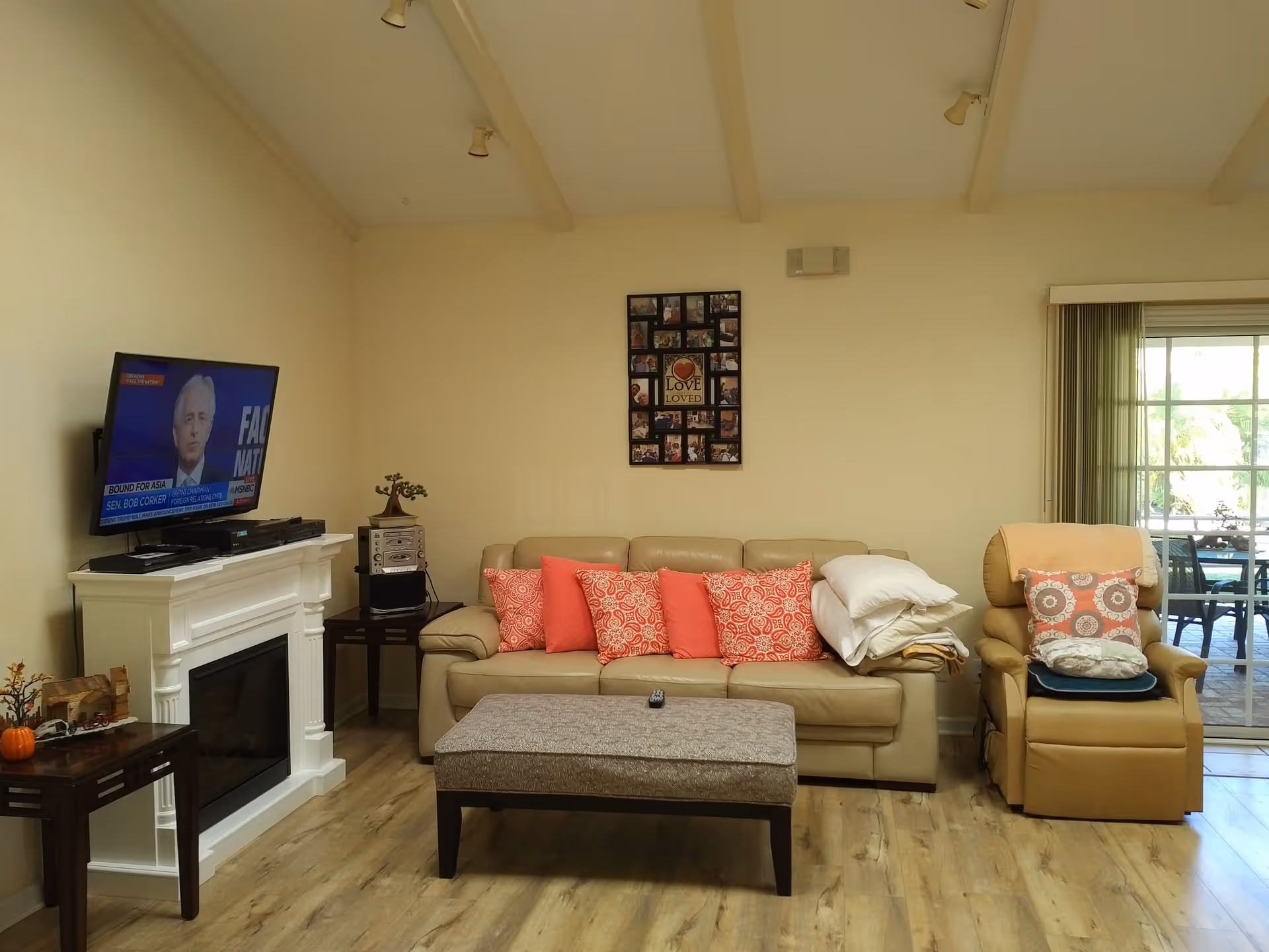 A cozy living room with a beige leather sofa adorned with coral and patterned pillows, a matching armchair with pillows and a blanket, a rectangular ottoman in front of the sofa, a white fireplace with a TV mounted above it showing a news program, and a sliding glass door leading to an outdoor patio.