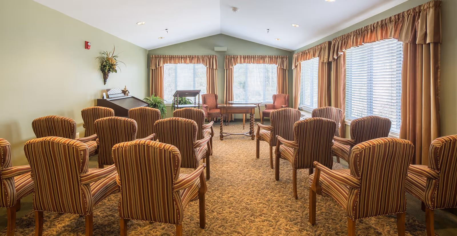 A well-lit room with multiple striped upholstered chairs arranged in rows facing a small wooden table and two armchairs near large windows with blinds and brown curtains. The walls are light green, and there is a decorative plant mounted on the wall and a small wooden cabinet in the corner.