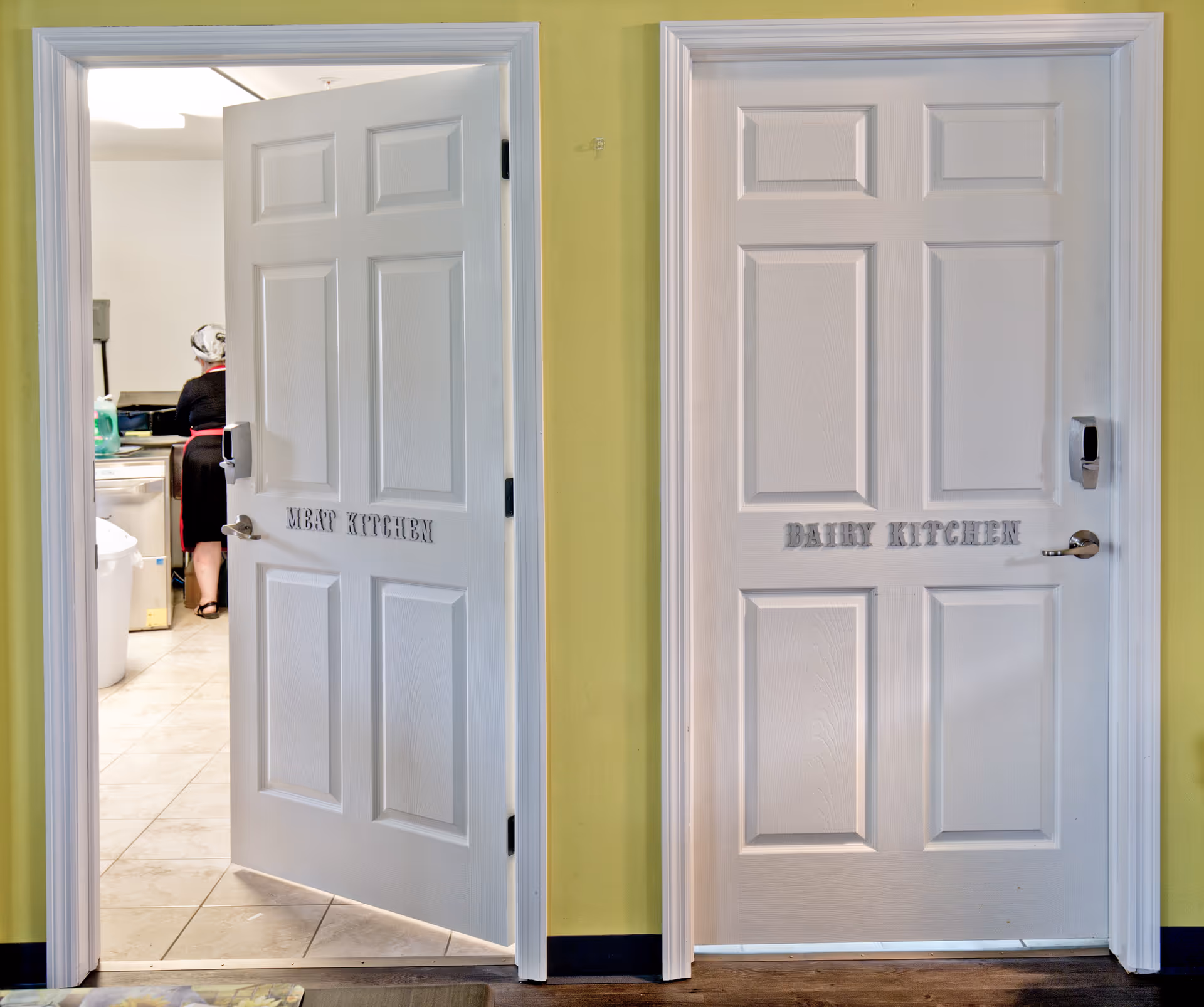 Two white doors labeled 'MEAT KITCHEN' and 'DAIRY KITCHEN' on a yellow wall. The door to the meat kitchen is open, showing a person working inside a kitchen area with tiled floor.