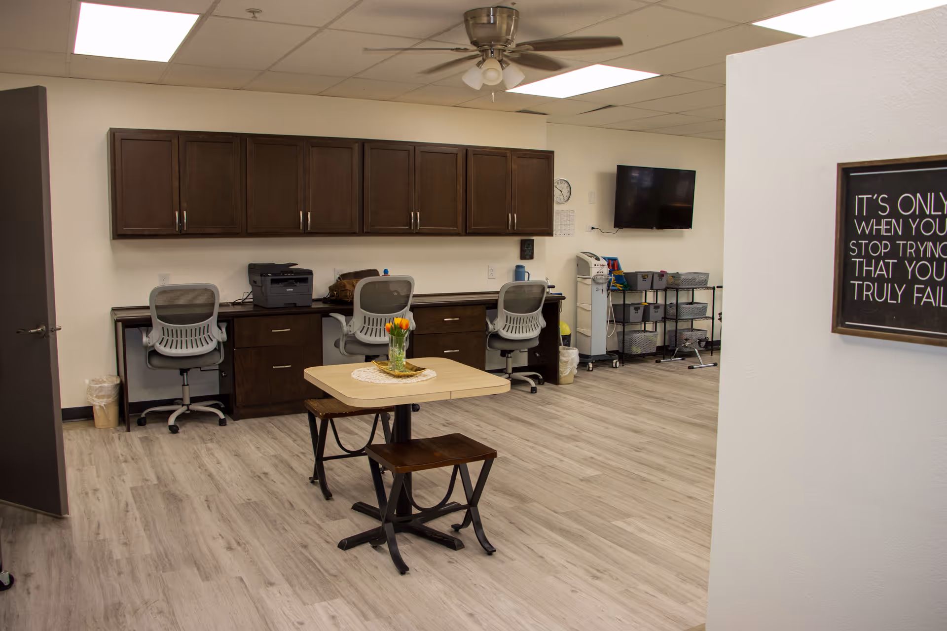 A clean and organized office or work area with three gray office chairs in front of a long counter with dark wooden cabinets above and below. A small table with two benches and a vase of orange tulips is in the center of the room. The floor is light wood, and there is a ceiling fan with lights. A wall-mounted TV and storage bins on a metal rack are visible in the background. A motivational framed sign is partially visible on the right wall.