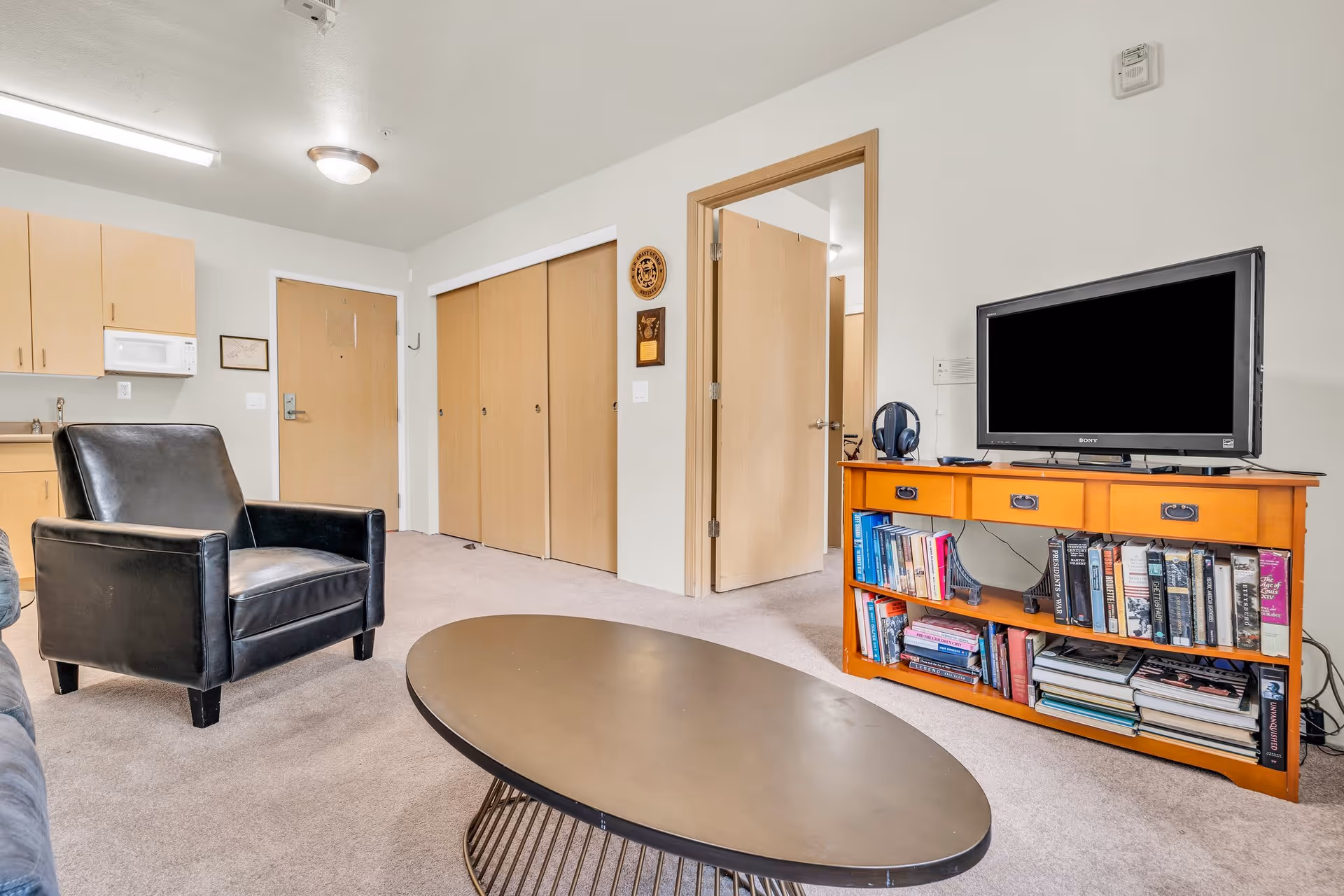 Interior view of a senior living facility room featuring a black leather armchair, an oval coffee table with a metal base, a wooden TV stand filled with books and a flat-screen TV on top. The room has beige carpet, light-colored walls, a kitchenette area with cabinets and a microwave, and multiple wooden doors including a closet and an open doorway leading to another room.