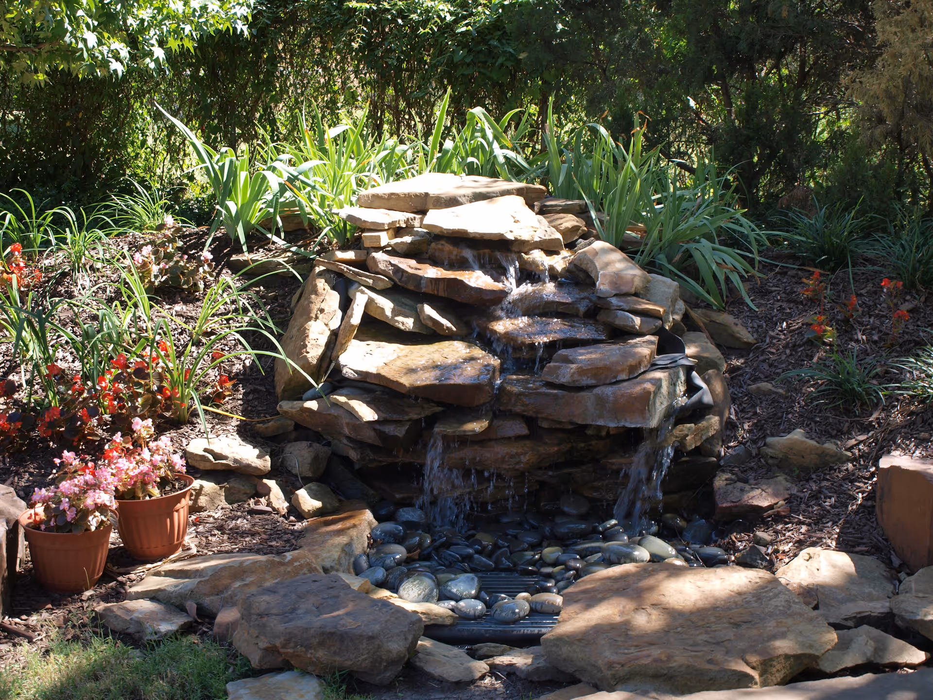 A small multi-tiered rock waterfall fountain with water flowing over stones, surrounded by garden plants and potted flowers.