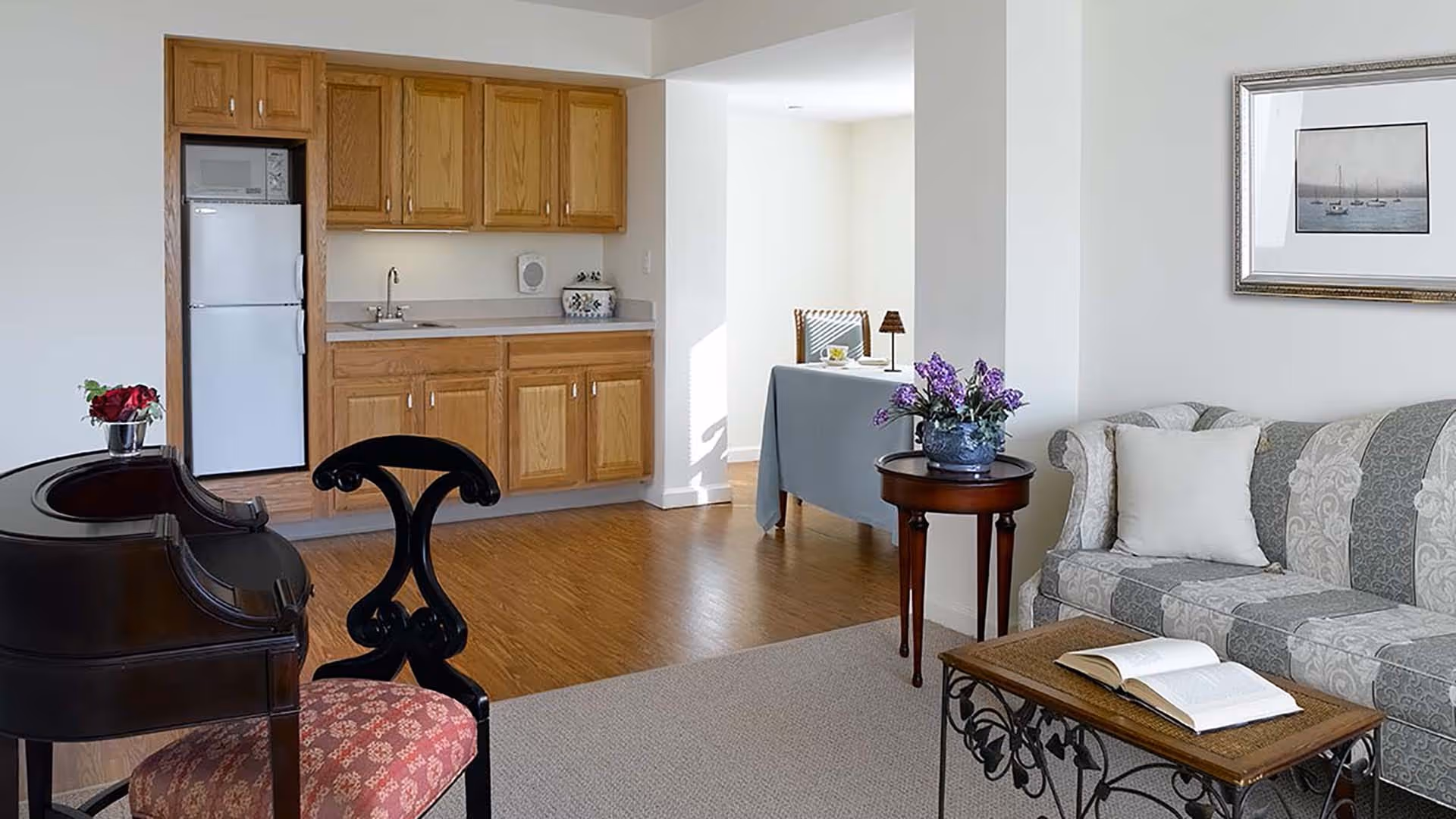 A cozy living area with a patterned sofa, a wooden coffee table with an open book, a small round side table with a vase of purple flowers, and a black wooden chair with a red cushioned seat. In the background, there is a kitchenette with wooden cabinets, a white refrigerator, and a microwave. A small dining table with a chair and a lamp is visible in an adjacent room.