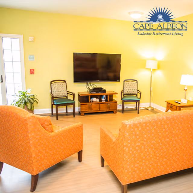 A cozy living room with two orange patterned armchairs facing a wall-mounted flat screen TV. Below the TV is a wooden media console with electronic devices and books. Two wooden chairs with patterned cushions are placed on either side of the console. The room has yellow walls, a floor lamp, a table lamp on a side table, and a potted plant near a glass door.
