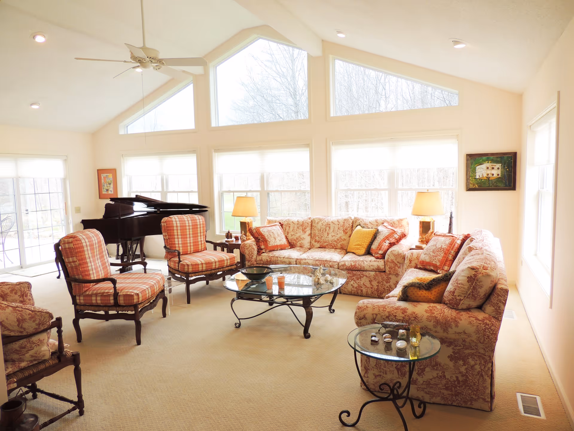 Bright living room with large triangular windows and multiple rectangular windows letting in natural light. The room features a floral patterned sofa set with matching cushions, two plaid armchairs, a glass coffee table, and a glass side table. There is a black grand piano in the corner near sliding glass doors leading outside. The walls are light-colored, and the ceiling has recessed lighting and a ceiling fan.