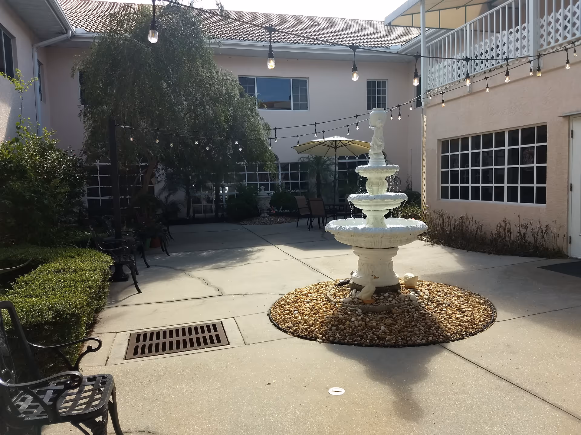 Sunlit courtyard with a three-tier white fountain surrounded by pebbles, string lights overhead, benches, and building windows.