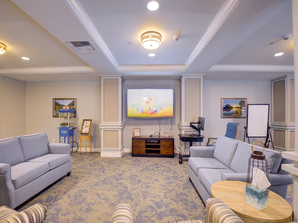 Well-lit communal living room with two sofas facing a wall-mounted TV, side tables, chairs, and a patterned carpet.