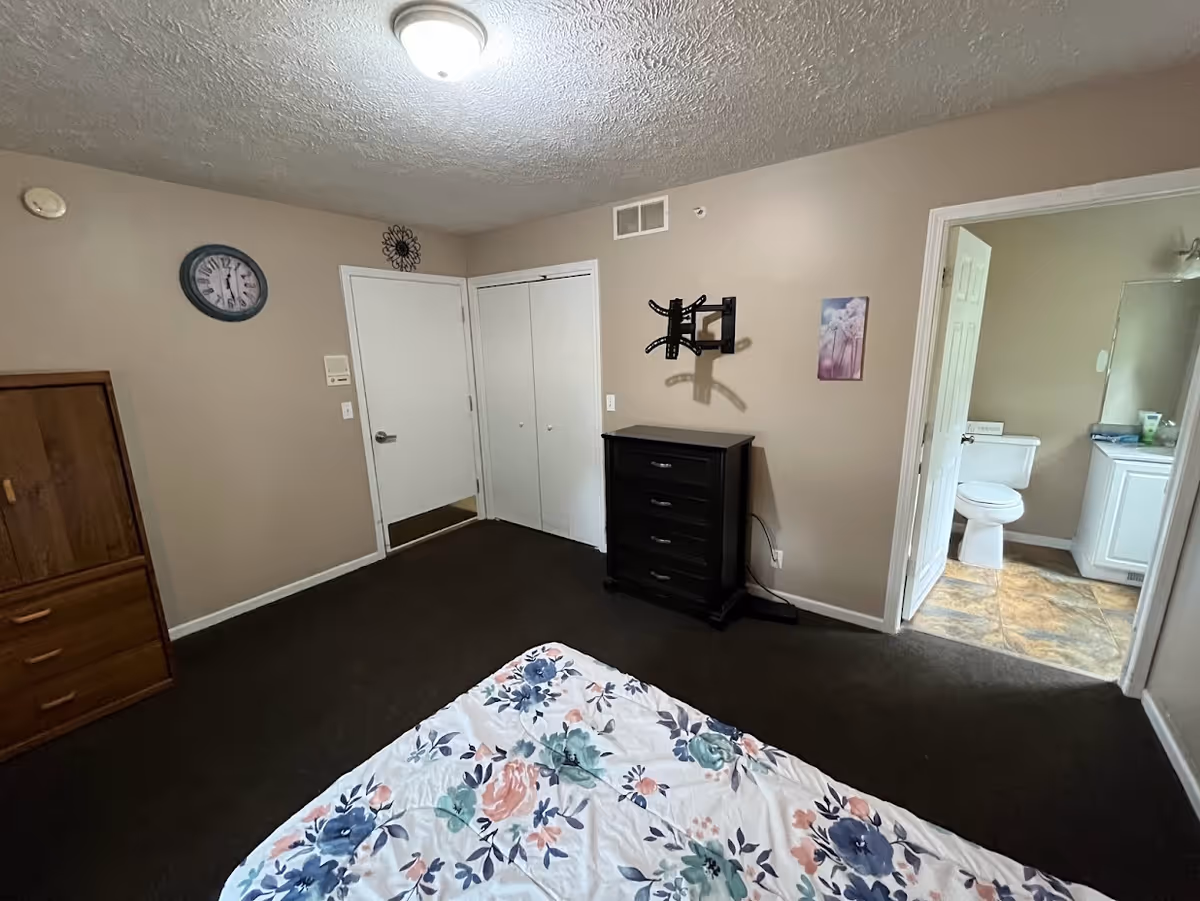 A bedroom with beige walls and dark carpet. There is a bed with a floral patterned bedspread in the foreground. Against the walls are a wooden dresser, a black chest of drawers, and a wall mount for a TV. A door leads to a bathroom with a toilet and sink visible. Another door and a closet with double doors are also visible. A clock and a decorative wall piece hang on the walls.