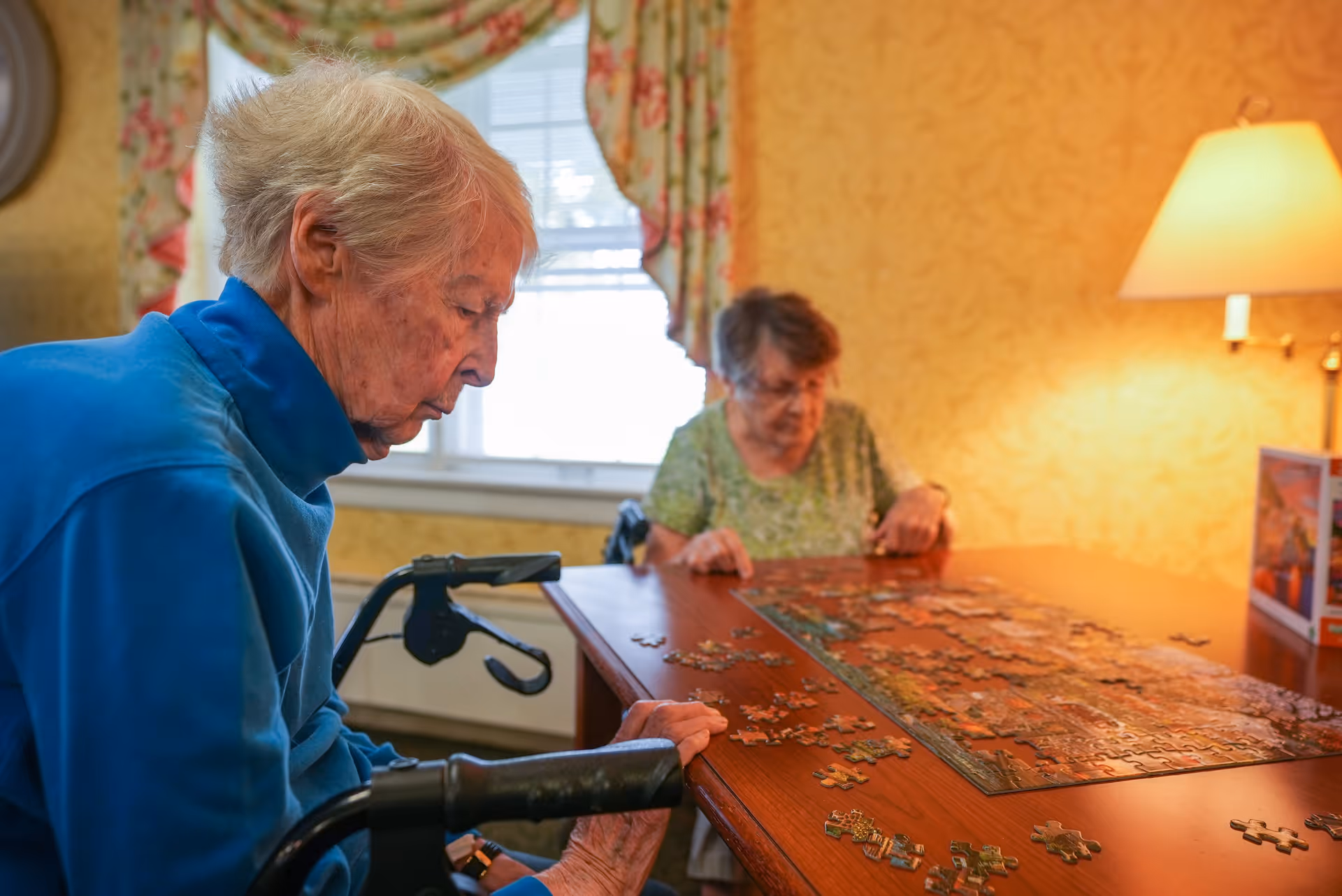 Two elderly women sitting at a wooden table working on a jigsaw puzzle together in a warmly lit room with floral curtains and a table lamp.