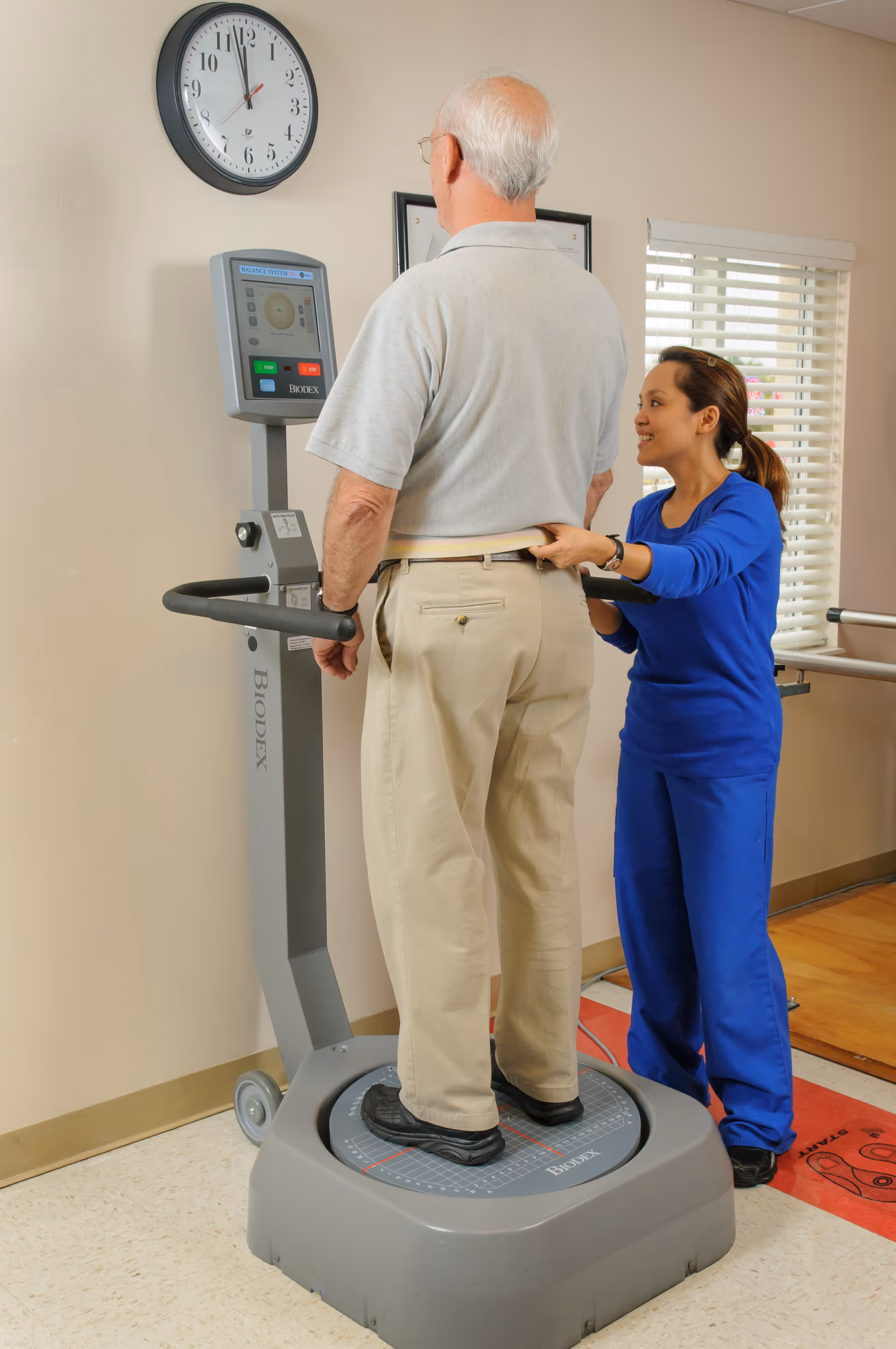 An elderly man stands on a Biodex balance testing machine while a healthcare professional in blue scrubs assists him by holding a belt around his waist. They are in a well-lit room with a clock on the wall showing 11:55 and a window with blinds.