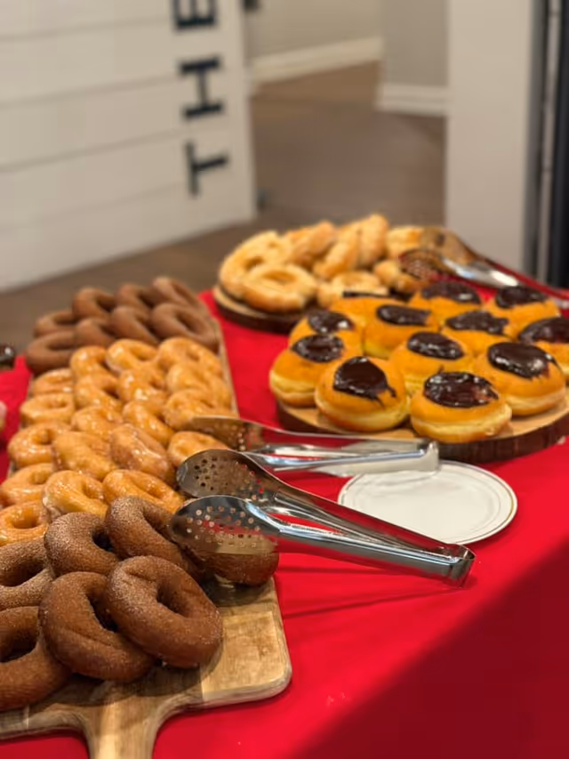 A variety of donuts displayed on wooden trays on a red tablecloth. The donuts include chocolate frosted, powdered sugar, glazed, and cinnamon sugar varieties. Metal tongs are placed on the trays for serving.