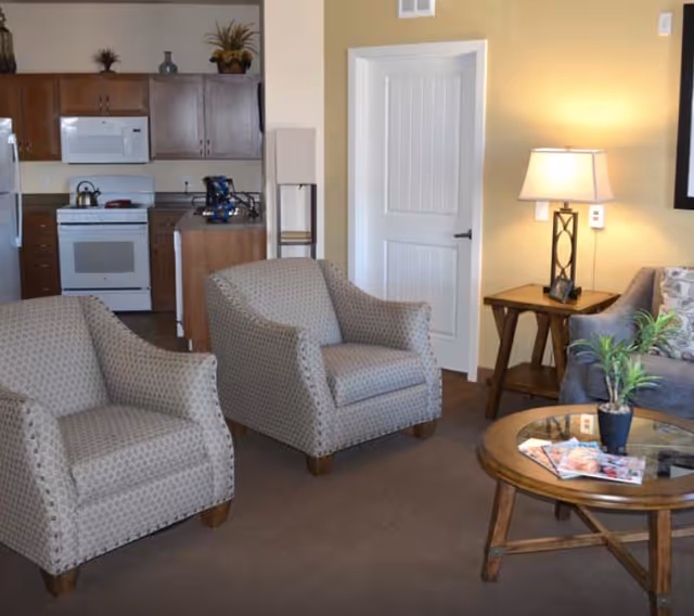 A cozy living area with two patterned armchairs, a round wooden coffee table with magazines and a small plant, a side table with a lamp, and a partial view of a kitchen with wooden cabinets and a white stove in the background.