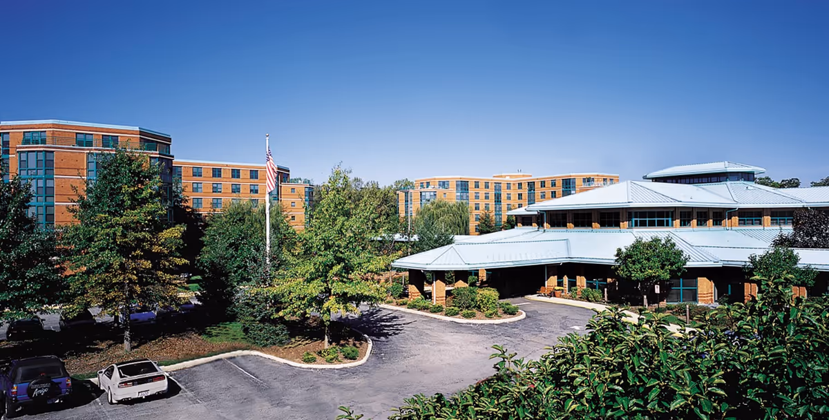 Exterior view of Ascension Living Bethlehem Woods facility showing multiple connected brick buildings with large windows, surrounded by trees and greenery under a clear blue sky. There is a driveway and parking area with a few cars, and an American flag on a flagpole near the entrance.