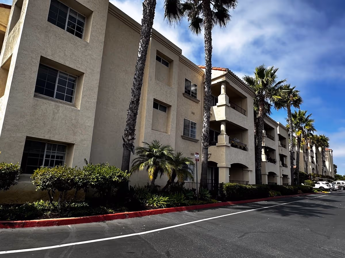 Three-story beige stucco building with balconies, palm trees, and a parking lane under a partly cloudy sky.