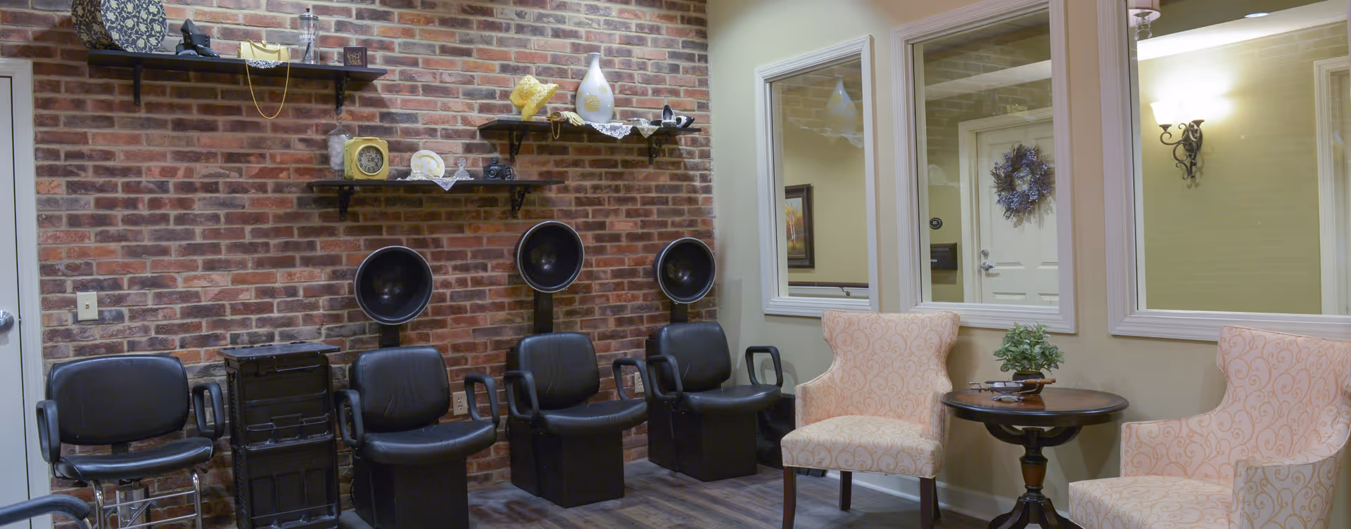 Interior salon area with three hooded hair dryers and black chairs against a brick wall, and two upholstered armchairs with a small table by windows.
