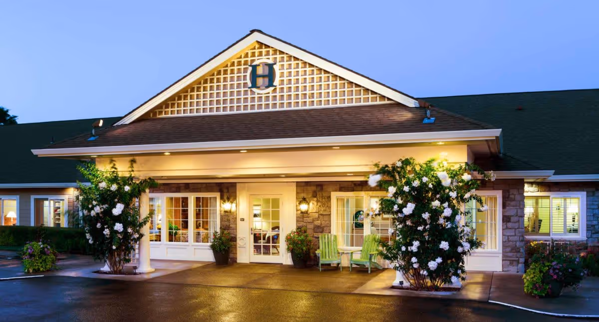 Exterior front entrance of The Hampton and Ashley Inn Memory Care Community building at dusk, featuring a covered porch with white columns, two green chairs with a small table, potted plants, and blooming white flowers on bushes near the entrance.