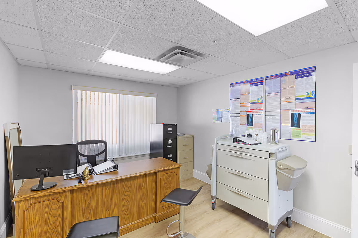 A small office room with a wooden desk, a computer monitor, two black chairs, a filing cabinet, and a medical cart with drawers. The walls are white with informational posters, and there is a window with vertical blinds letting in natural light.