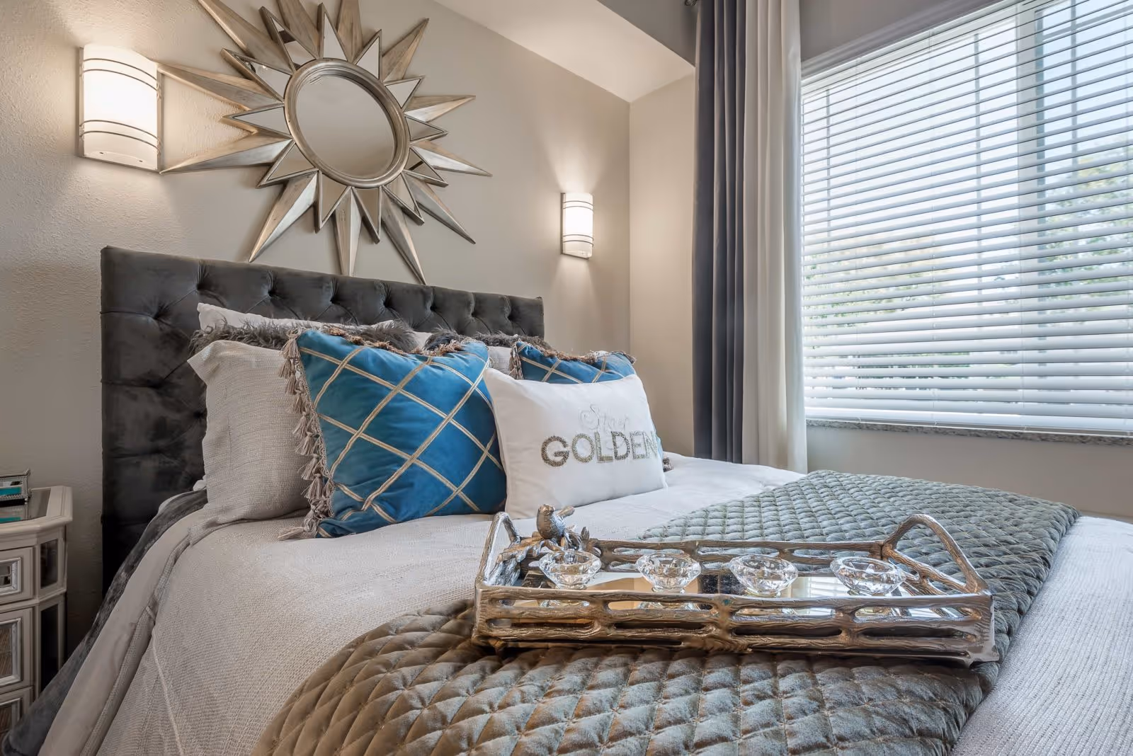 Sunlit bedroom with a tufted dark headboard, layered pillows and a decorative tray on the bed in front of a large window.