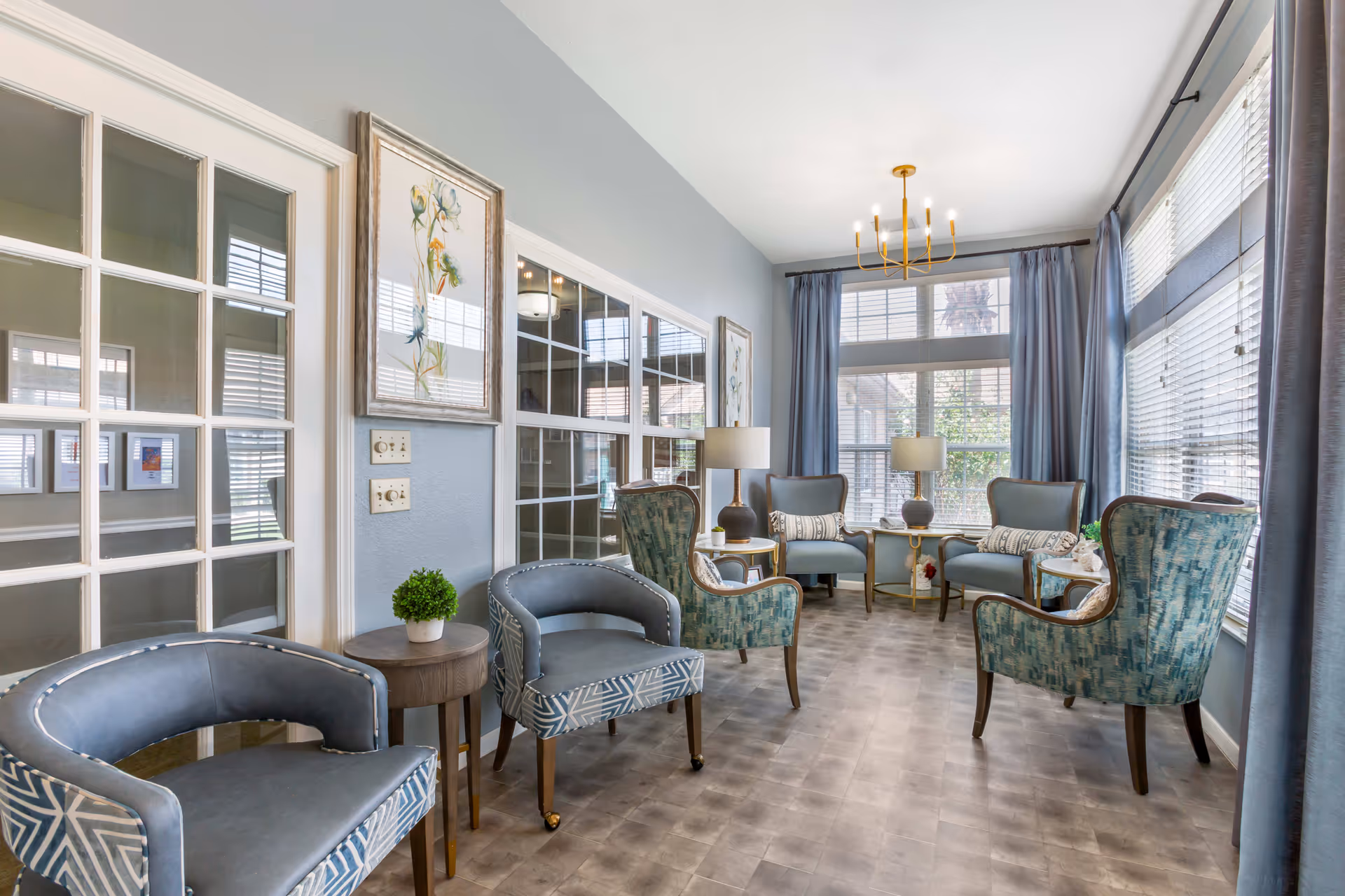 A bright and airy living room with multiple upholstered chairs arranged around small side tables. The room features large windows with blinds and blue curtains, light gray walls, and a modern gold chandelier hanging from the ceiling. There are framed floral artworks on the walls and two table lamps on side tables near the windows.