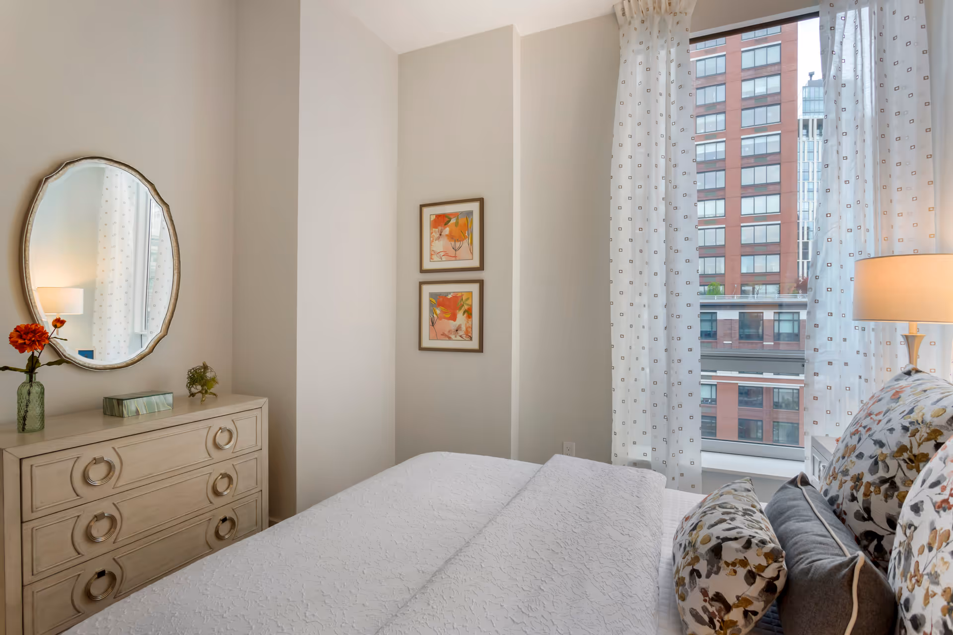 A cozy bedroom with a neatly made bed featuring floral patterned pillows and a white textured bedspread. To the left, there is a light-colored dresser with a round mirror above it, decorated with a small vase holding orange flowers and a box of tissues. Two framed floral artworks hang on the wall near the corner. Large windows with sheer white curtains allow natural light to brighten the room and offer a view of a tall brick building outside. A table lamp with a beige shade is placed on a bedside table next to the bed.